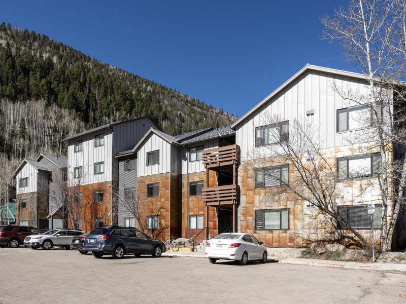 Modern three-story apartment building with metal and wood siding, parked cars in front, and a forested hillside in the background under a clear blue sky.