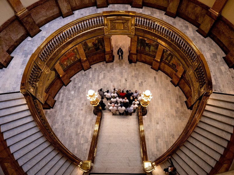 A group of people gathered at the base of a grand staircase in the ornate, circular hall of the Colorado State Capitol building, seen from above.