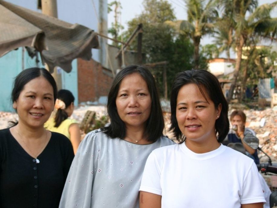 Three women stand outdoors in front of a street scene with debris, palm trees, and buildings in the background.