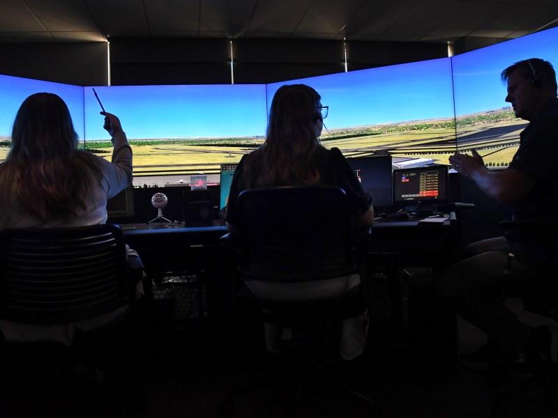 Three people sit at desks with monitors, facing a large panoramic flight simulator screen showing an airport runway scene.
