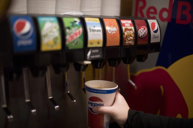 A person fills a Pepsi cup at a soda fountain machine with various drink options visible, including Pepsi, Mountain Dew, and Dr Pepper.