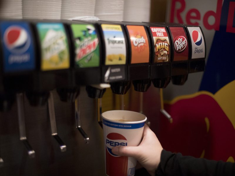 A person fills a Pepsi cup at a soda fountain machine with various drink options visible, including Pepsi, Mountain Dew, and Dr Pepper.