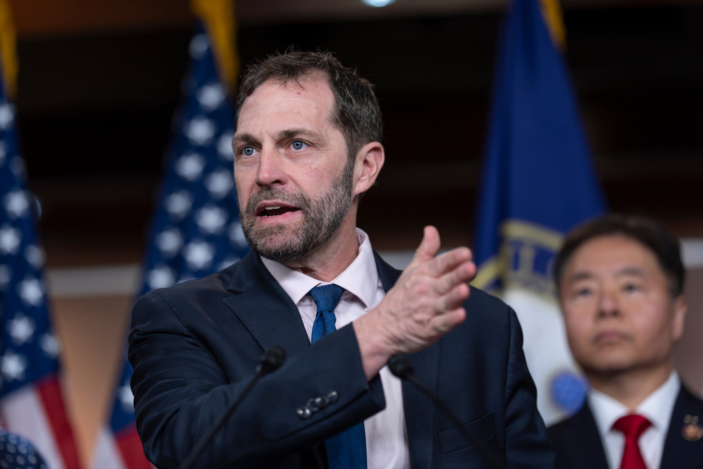A photo of U.S. Rep. Jason Crow. He is gesturing with his left hand. He wears a dark suit, white shirt and blue tie. There is a man in the background wearing a red tie.