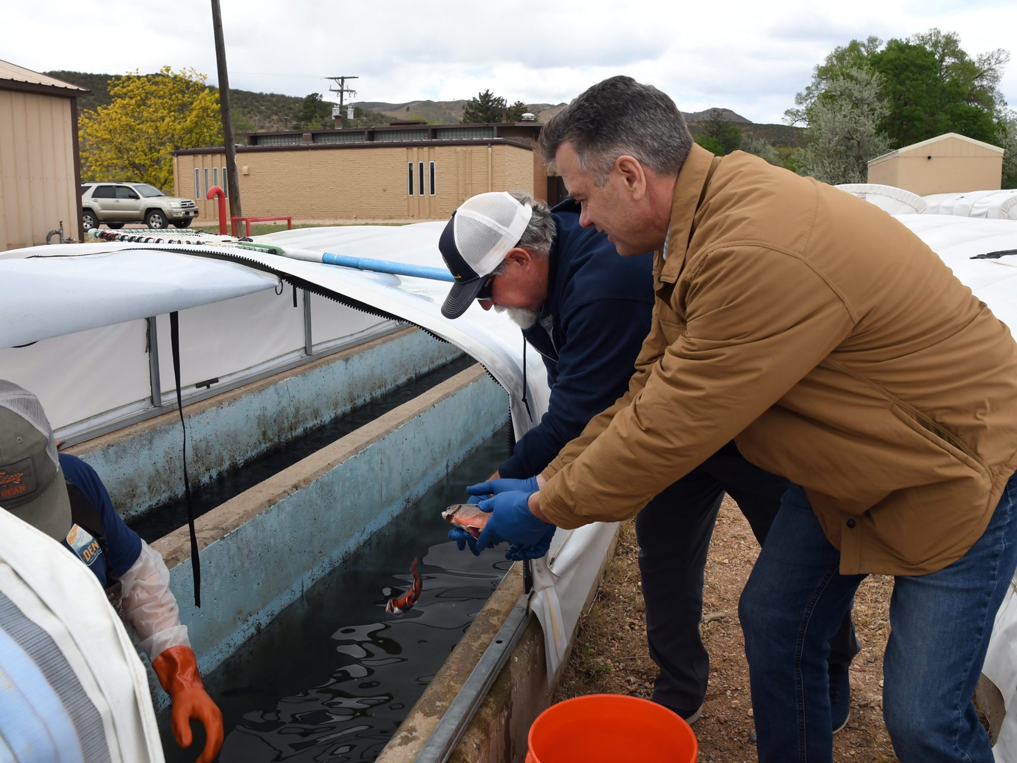 Two men examine trout at an outdoor aquaculture facility, standing by a water tank under a white canopy. A third person is partially visible on the left, wearing orange gloves.