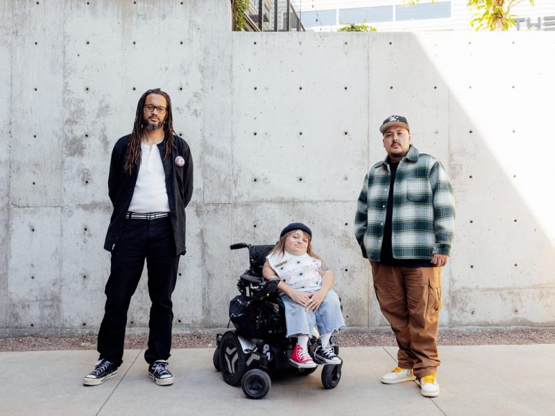 Three people pose in front of a concrete wall; one person uses a powered wheelchair, while the other two stand on either side.