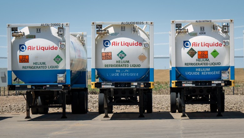 Three Air Liquide tanker trailers labeled “Helium, Refrigerated Liquid” are parked side by side on a paved surface near a chain-link fence.