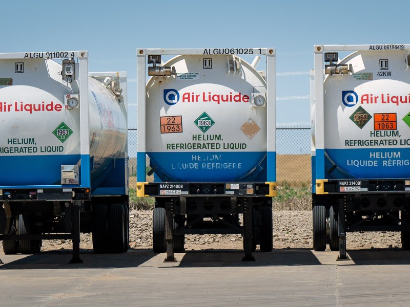 Three Air Liquide tanker trailers labeled “Helium, Refrigerated Liquid” are parked side by side on a paved surface near a chain-link fence.