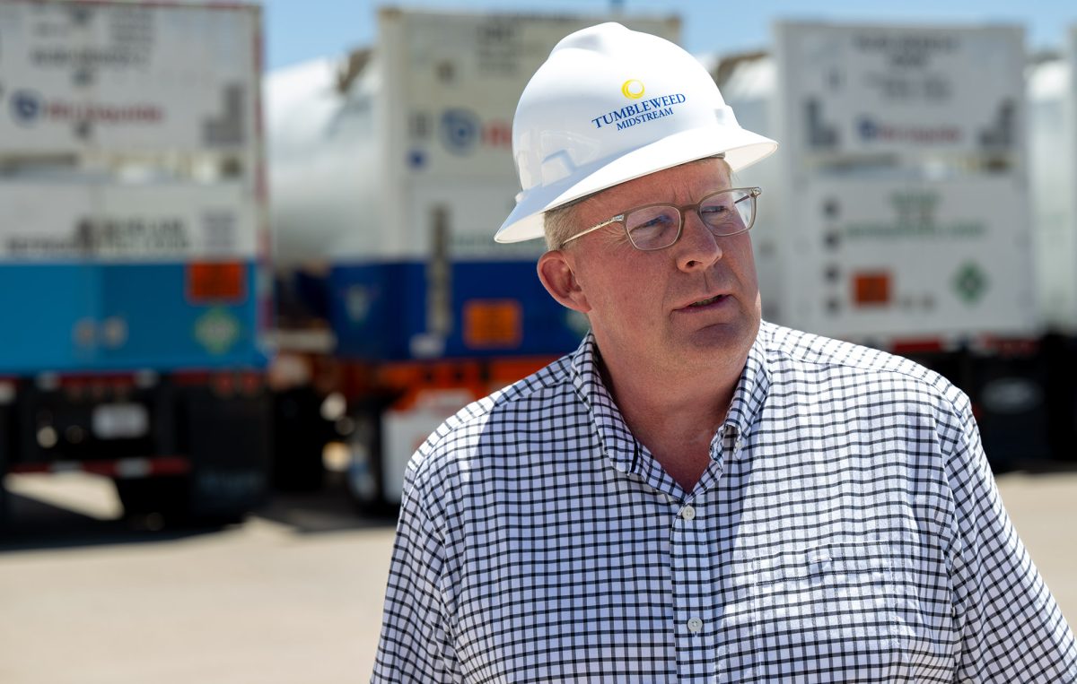 A man wearing glasses and a white hard hat stands outdoors near tankers specially designed to transport liquefied helium