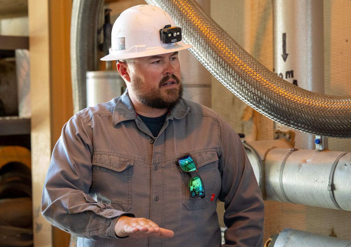 A man wearing a white hard hat with a camera affixed to it stands in front of equipment used to liquefy helium