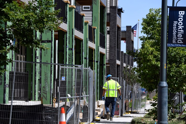 A man wearing a yellow safety shirt walks near fencing in front of townhomes that are under construction.