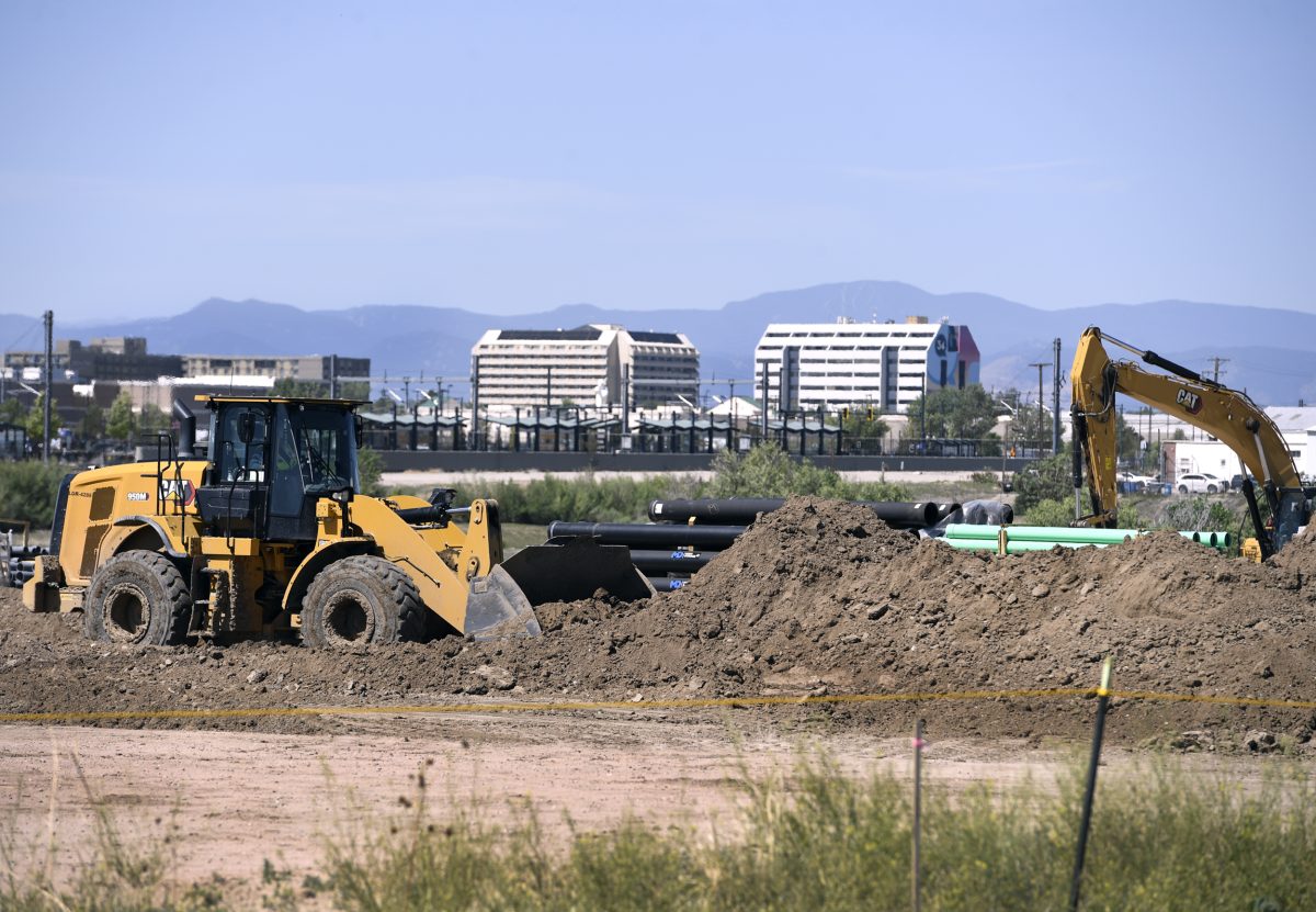 A bulldozer and an excavator move dirt at a construction site with pipes in the foreground and office buildings and mountains in the background.