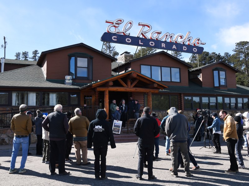 A group of people and media gather outside El Rancho Colorado, a rustic-style building with a large sign, on a sunny day.