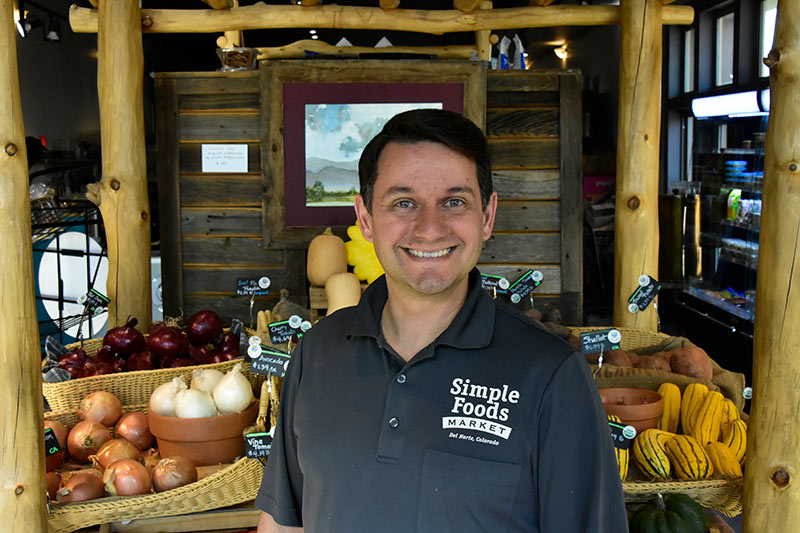A man wearing a "Simple Foods Market" polo shirt stands and smiles in front of a produce display with onions, squash, and other vegetables.