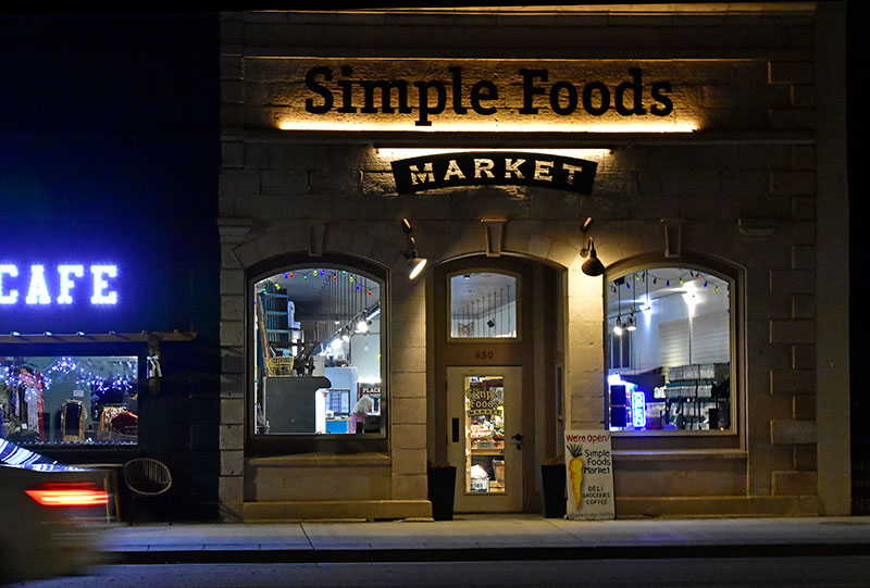 Nighttime view of Simple Foods Market storefront lit by exterior lights, with a partially visible illuminated café sign to the left.