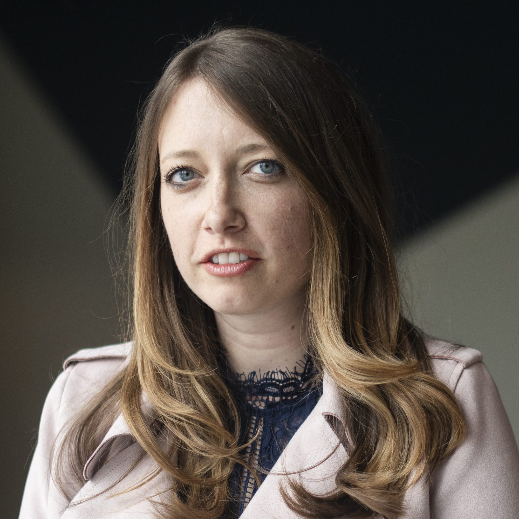 A woman with long hair wearing a light pink coat and a dark lace top looks towards the camera, standing against a dark background.