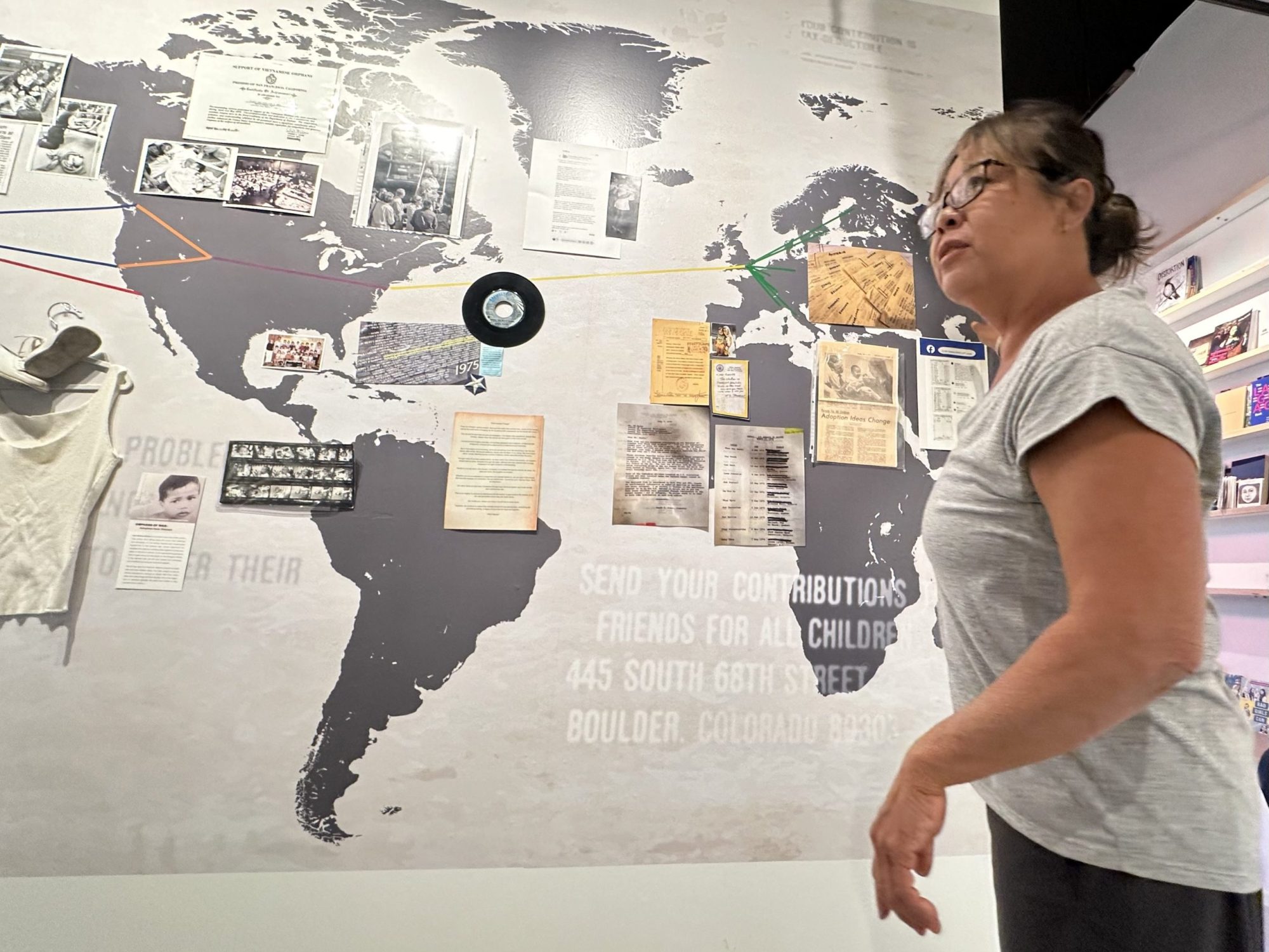 A woman stands in front of a wall display featuring a world map with photos, documents, and colored lines connecting various locations.