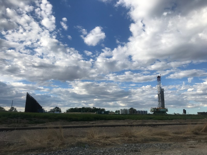 An oil and gas drilling rig is visible behind a barrier. The sky is blue and filled with puffy clouds
