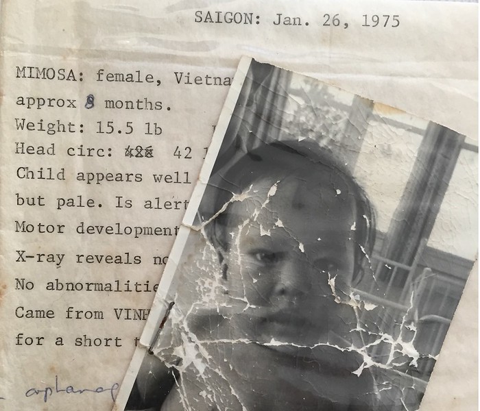 A worn black-and-white photo of a young child is placed over a typed medical report from Saigon dated January 26, 1975, detailing the child's health information.