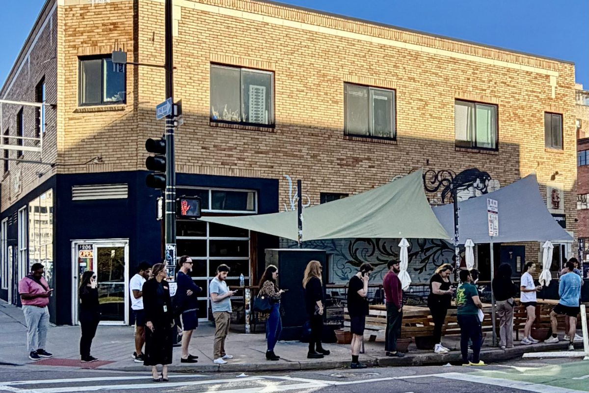 People stand in a line outside a corner brick building with large windows and a tented outdoor seating area on a sunny day.