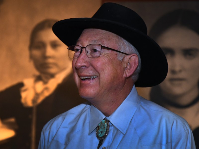 An older man in a black hat, glasses, and bolo tie smiles in front of two sepia-toned historical portraits.