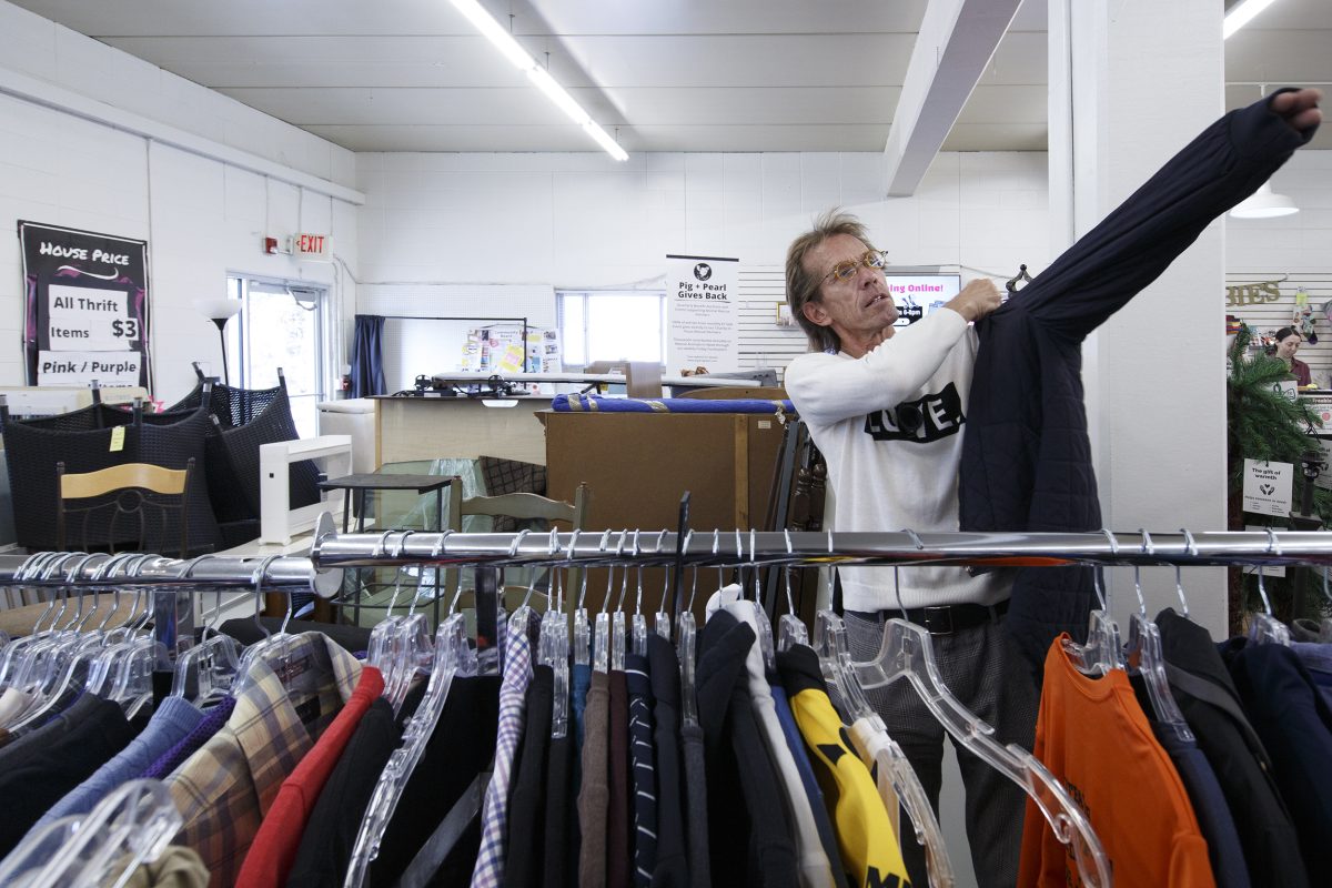 A person tries on a dark jacket in a thrift store, standing behind a rack of assorted clothing under fluorescent lighting.