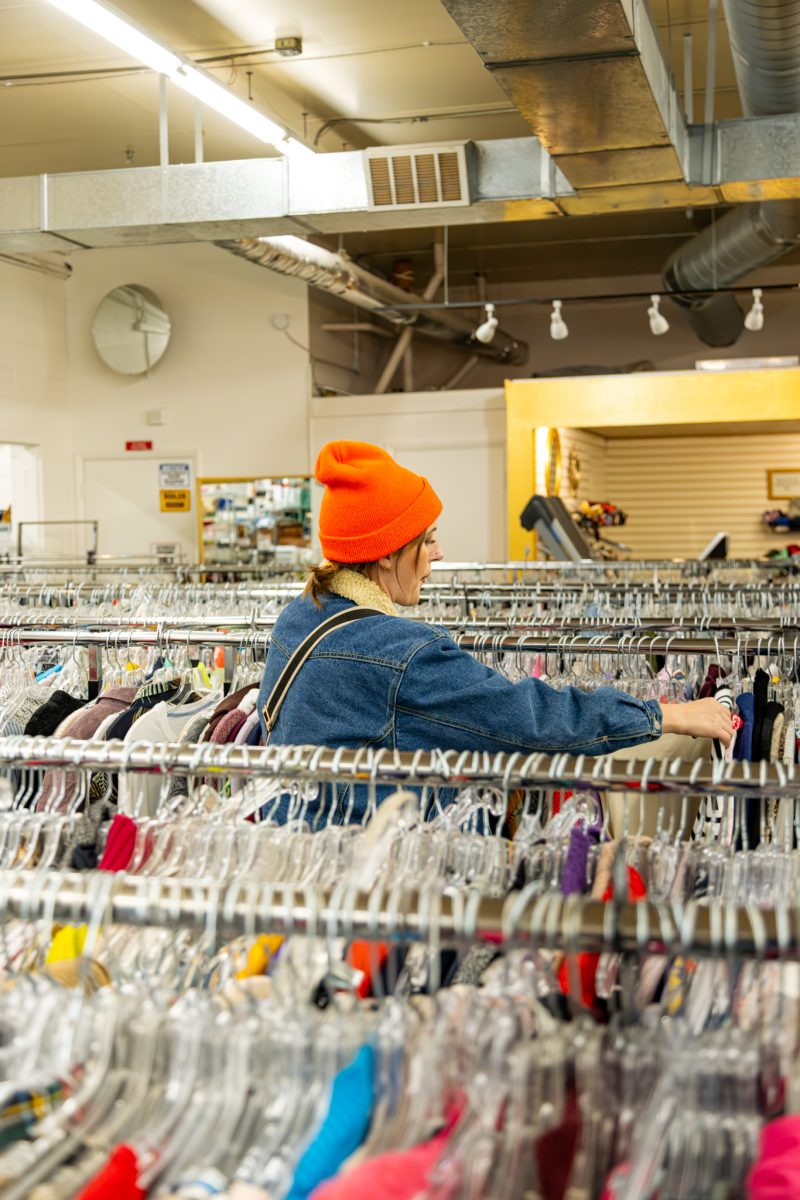 A person wearing an orange beanie and denim jacket browses clothing racks in a thrift store.