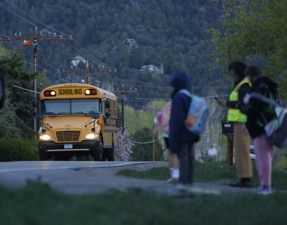 A yellow school bus approaches as children with backpacks wait at the side of a rural road in the early morning.