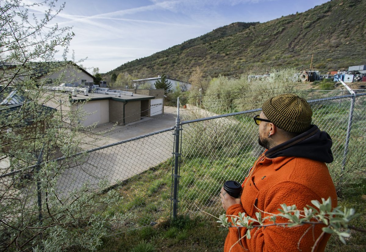 A person in an orange jacket holds a coffee cup while standing behind a chain-link fence, looking toward buildings and hills in the distance.