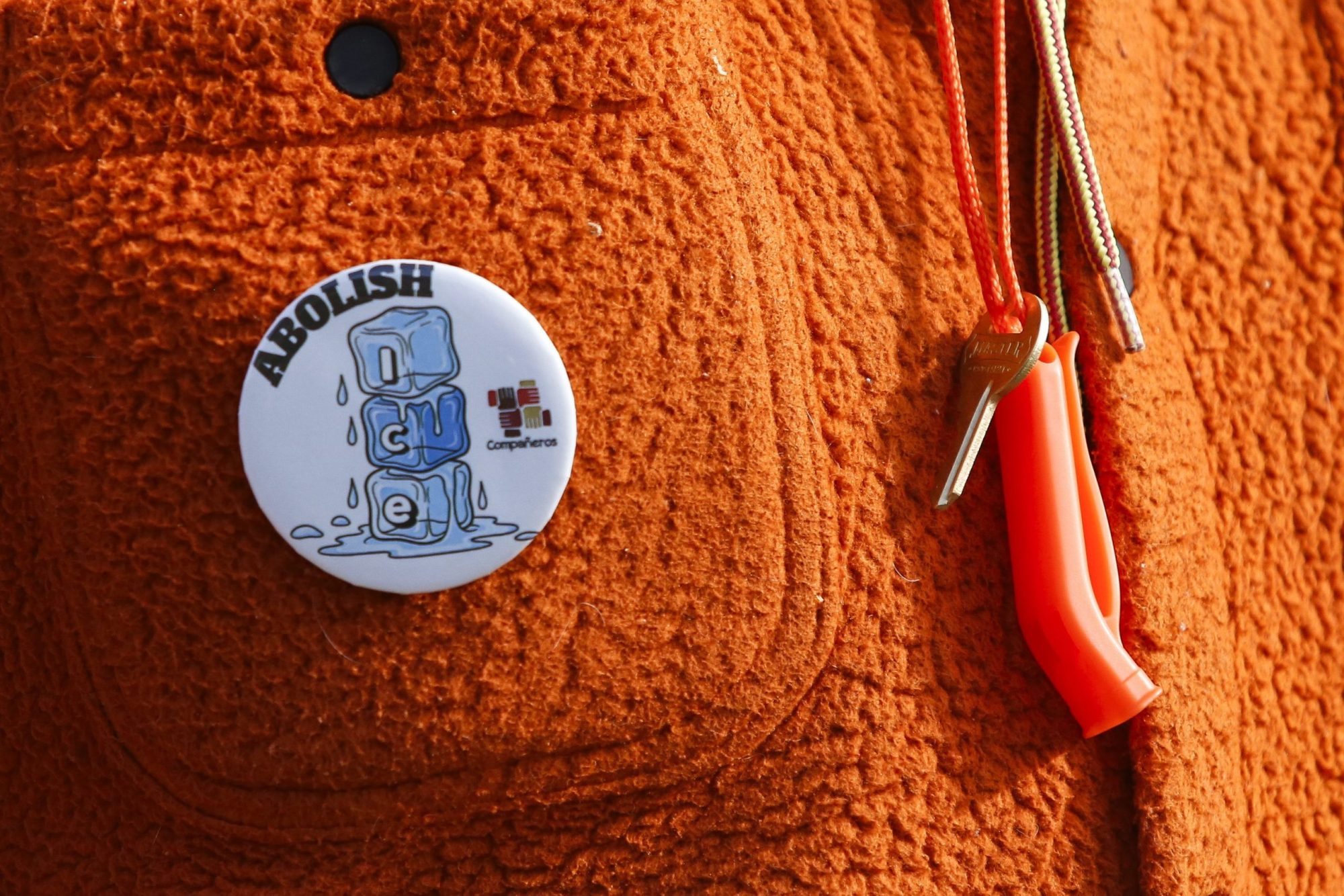 A white button pin reading "Abolish ICE" is attached to an orange textured jacket, next to two whistles on lanyards.