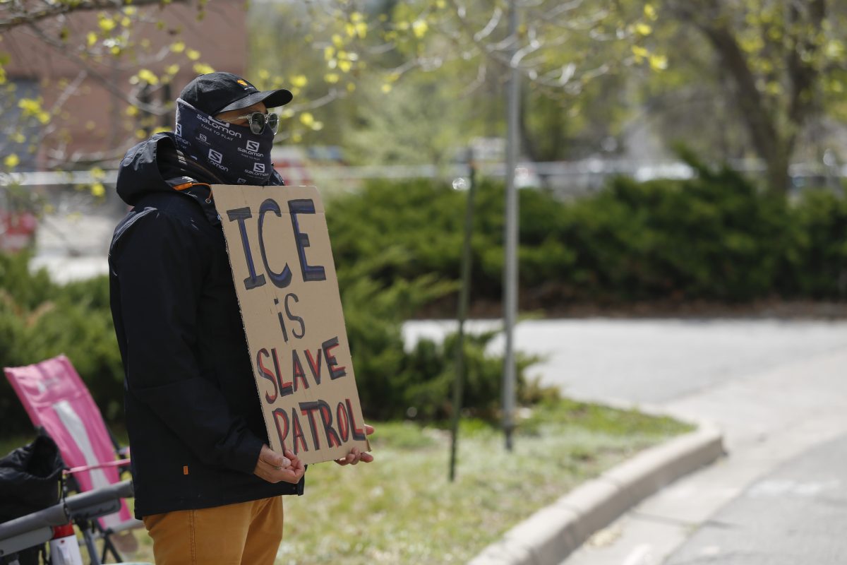 A person wearing a mask and hat holds a cardboard sign that reads "ICE is SLAVE PATROL" while standing outdoors near a street.