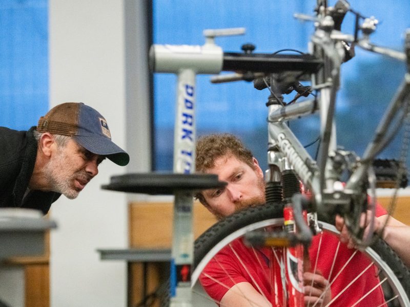 Two men closely examine and repair a bicycle in an indoor setting, with tools and a bike stand visible in the foreground.