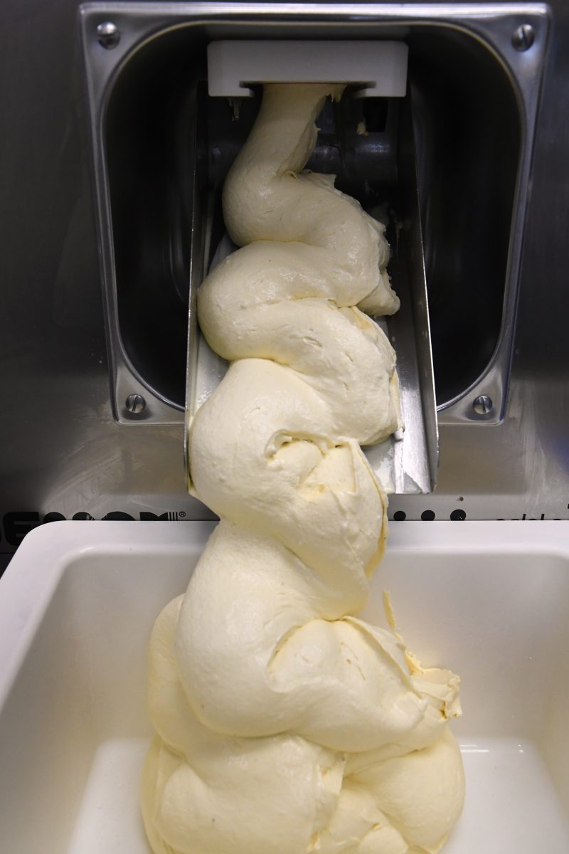 Thick, creamy gelato being dispensed from a commercial machine into a white container.