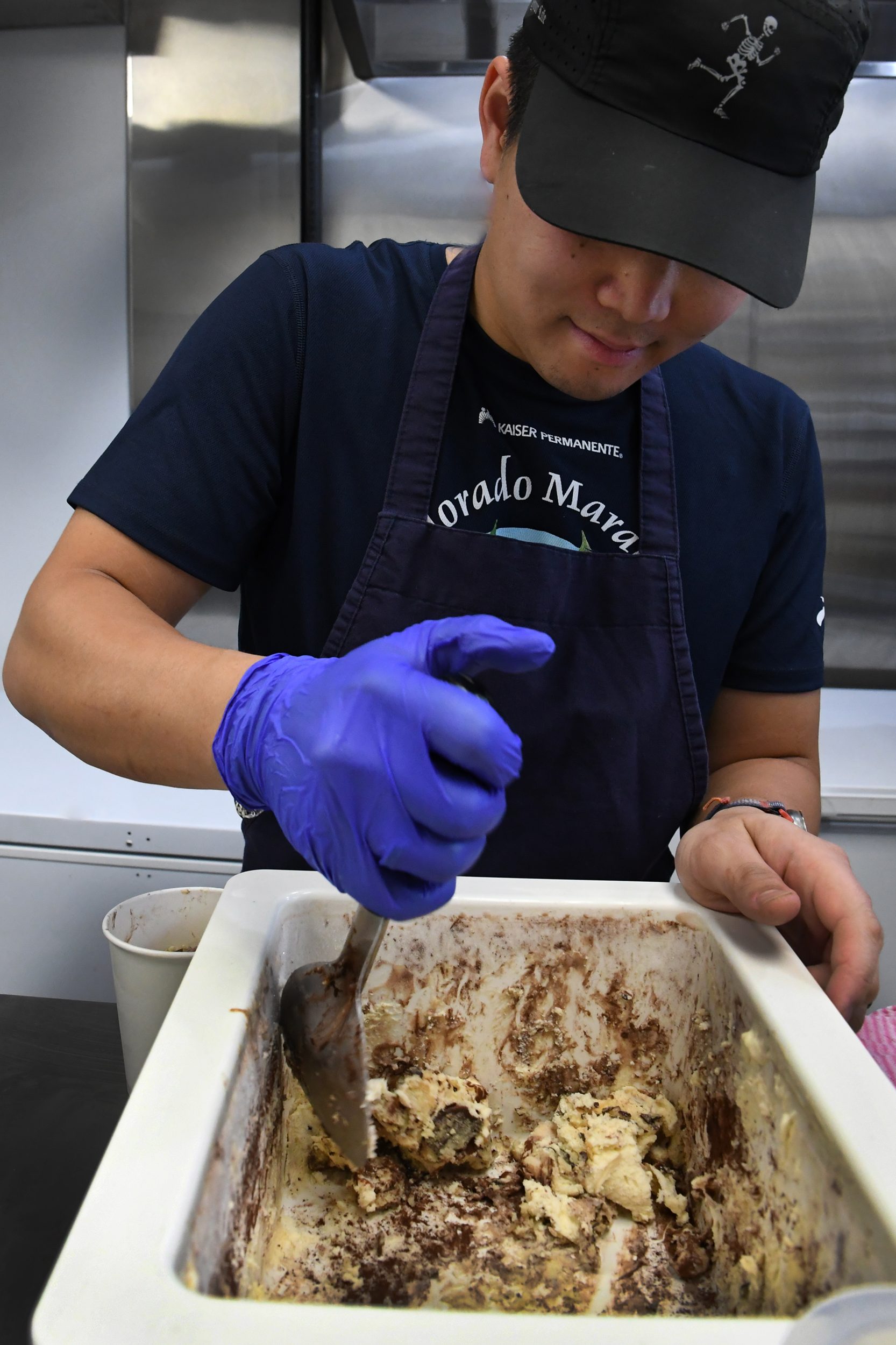 A person wearing a black cap, apron, and purple gloves mixes cookie dough in a white container with a metal utensil in a kitchen.