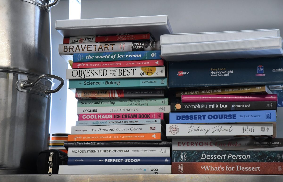 A stack of assorted baking and dessert cookbooks is arranged on a kitchen shelf next to a large metal container.