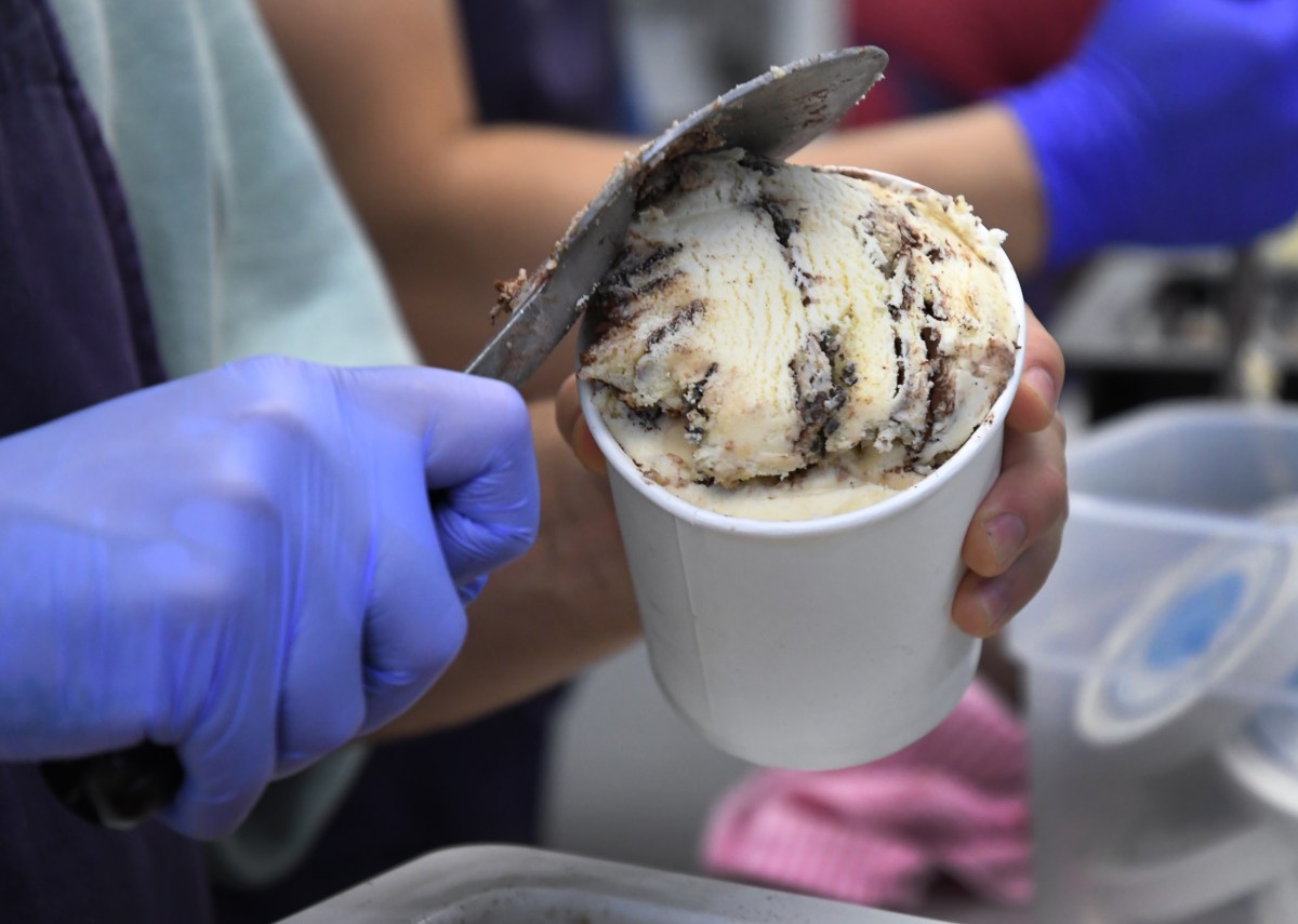 A person wearing blue gloves scoops ice cream with chocolate swirls into a white paper cup.