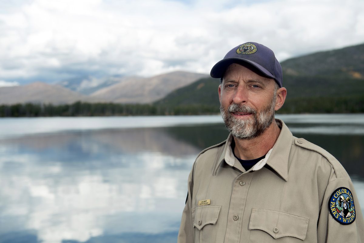 A park ranger in uniform stands by a lake with mountains and trees in the background under a cloudy sky.