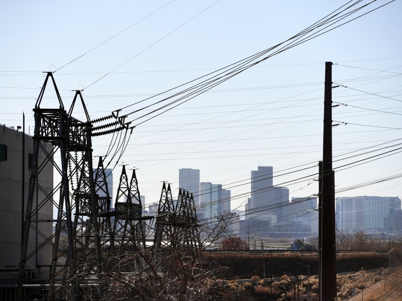 Xcel Energy power lines and electrical towers in the foreground with a city skyline in the distance under a clear sky.