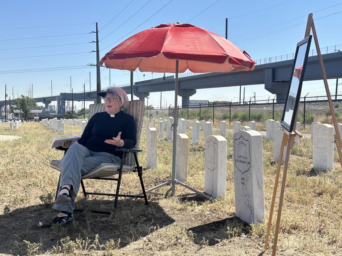 A woman wearing a black shirt and clerical collar sits in a lawn chair under a red umbrella next to the grave of Silas Soule.