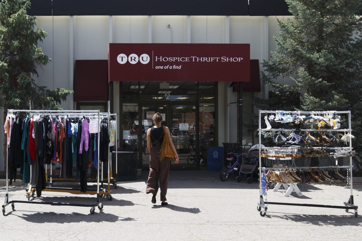 A person walks toward the entrance of a hospice thrift shop, with two clothing racks displaying garments and accessories outside the store.