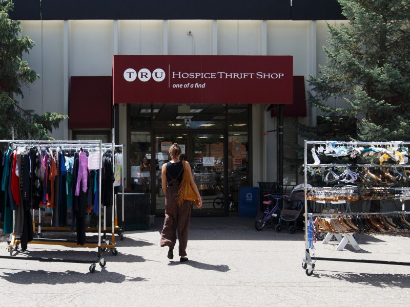 A person walks toward the entrance of a hospice thrift shop, with two clothing racks displaying garments and accessories outside the store.