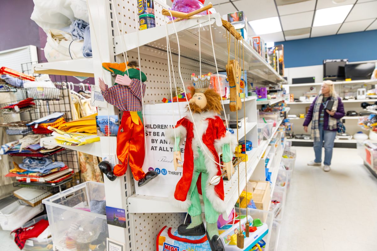Two marionette puppets hang from a shelf in a thrift store aisle filled with assorted items; a person walks in the background.