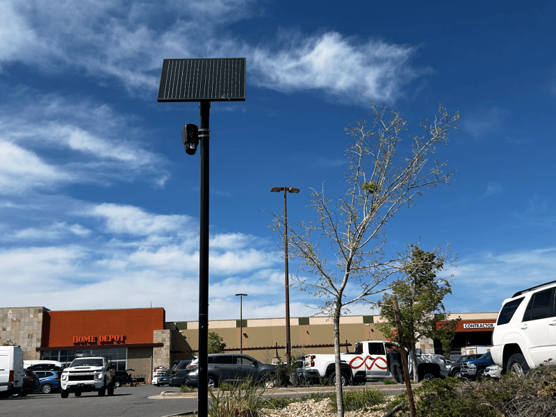 A solar-powered security camera is mounted on a pole in a parking lot outside a Home Depot store, with cars parked nearby under a blue sky with scattered clouds.