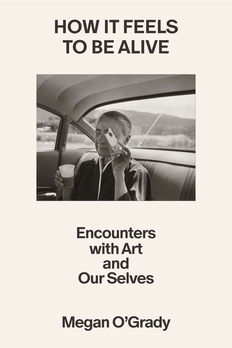 Book cover for "How It Feels to Be Alive" by Megan O’Grady, featuring a black-and-white photo of a person sitting in a car, holding a drink and a paper mask.