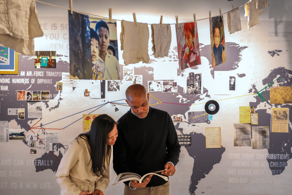 Two people look at a book in front of a world map display, with photographs, documents, and clothing hanging above them on a clothesline.