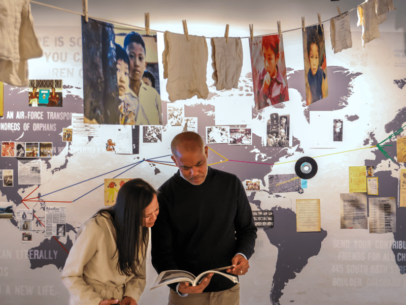 Two people look at a book in front of a world map display, with photographs, documents, and clothing hanging above them on a clothesline.