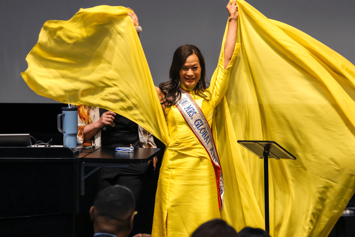 A woman in a bright yellow dress holds out the fabric, wearing a "Mrs. Global" sash, standing on a stage in front of an audience.