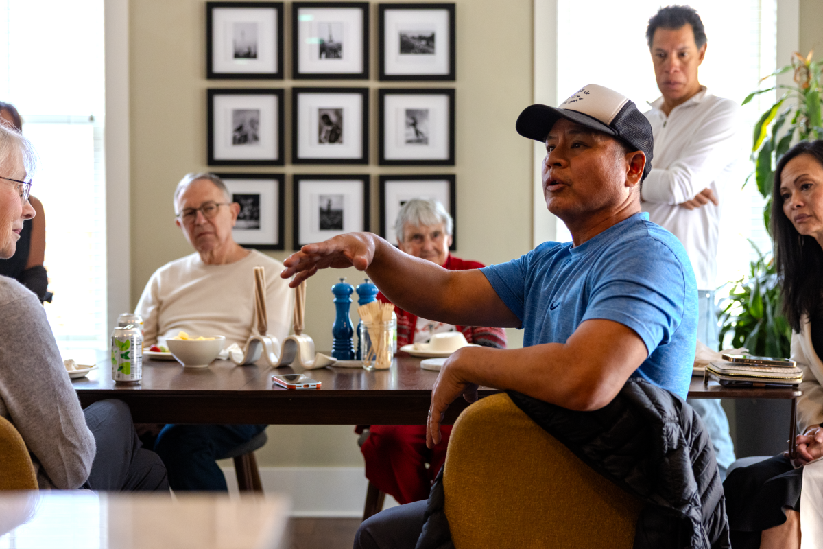A group of people sit around a dining table, engaged in discussion. One man in a blue shirt and cap gestures while talking, and others listen attentively.