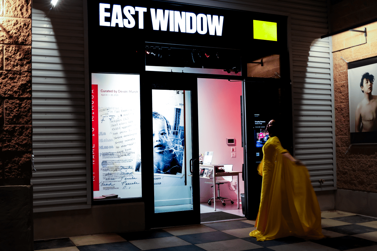 A person in a flowing yellow dress stands outside the illuminated entrance of a gallery called EAST WINDOW at night.
