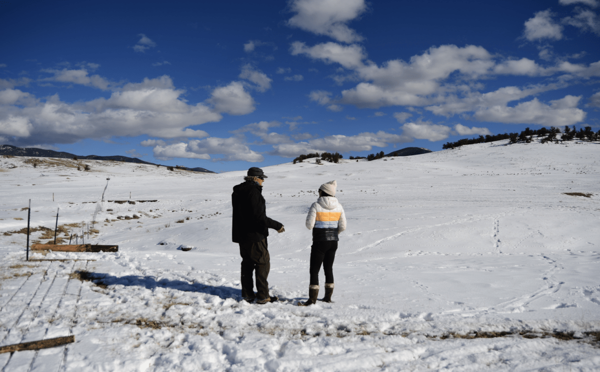 A woman in a beanie talks to a man in black while they stand on a snowy field, looking at elk tracks.
