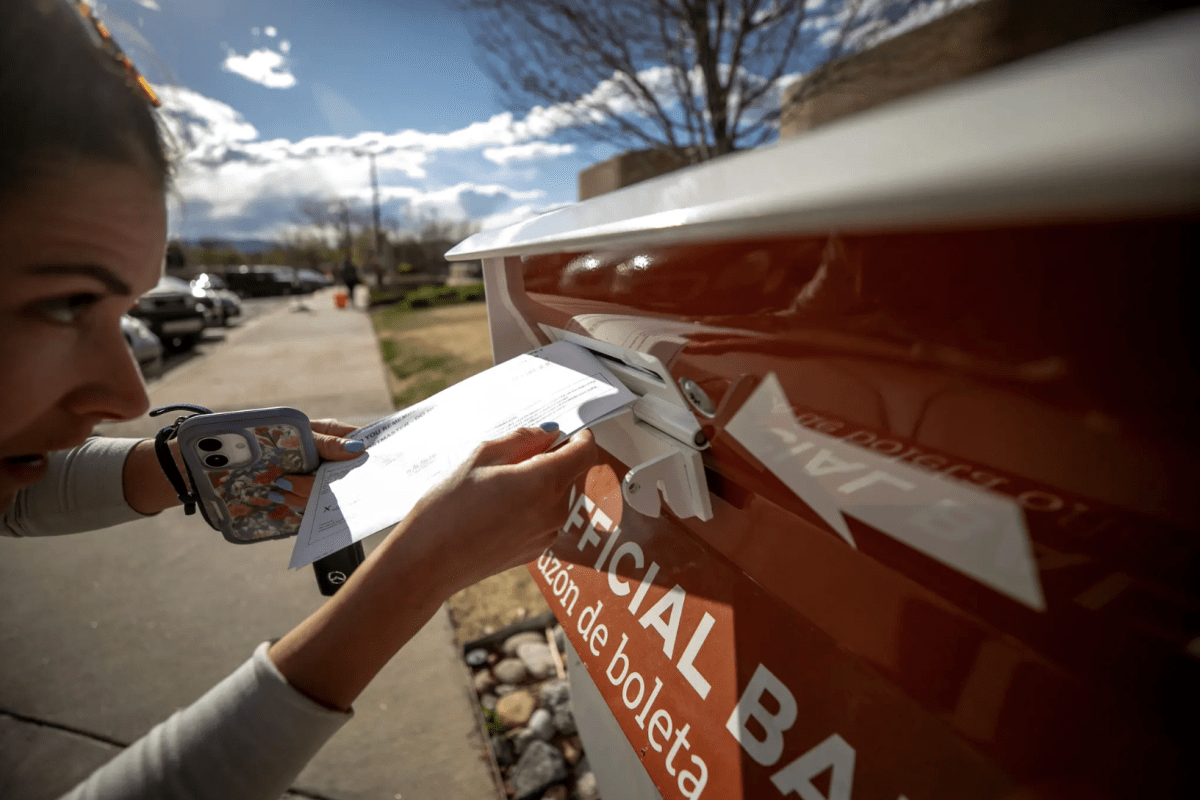 A woman places her ballot into a red ballot box. She is holding a cell phone in her hand.
